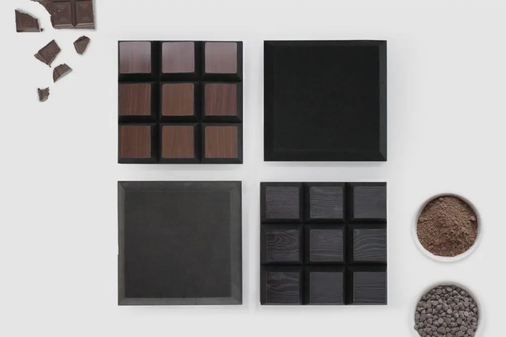Four square trays with chocolate bar designs and empty slots, surrounded by broken chocolate pieces, chocolate chips, and a bowl of cocoa powder, all arranged neatly on a white background.